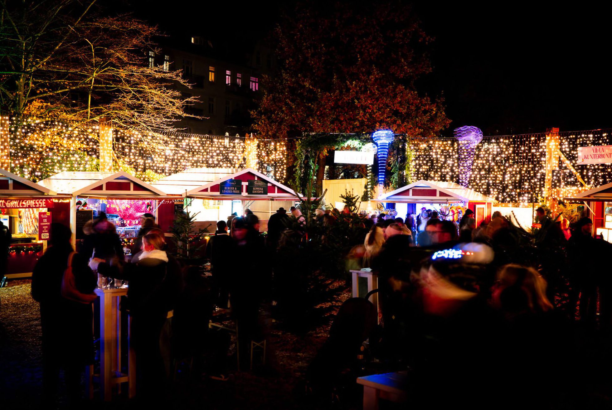 Ein Weihnachtsmarkt mit hell erleuchteten Ständen und Lichterketten, Besucher im Gespräch.