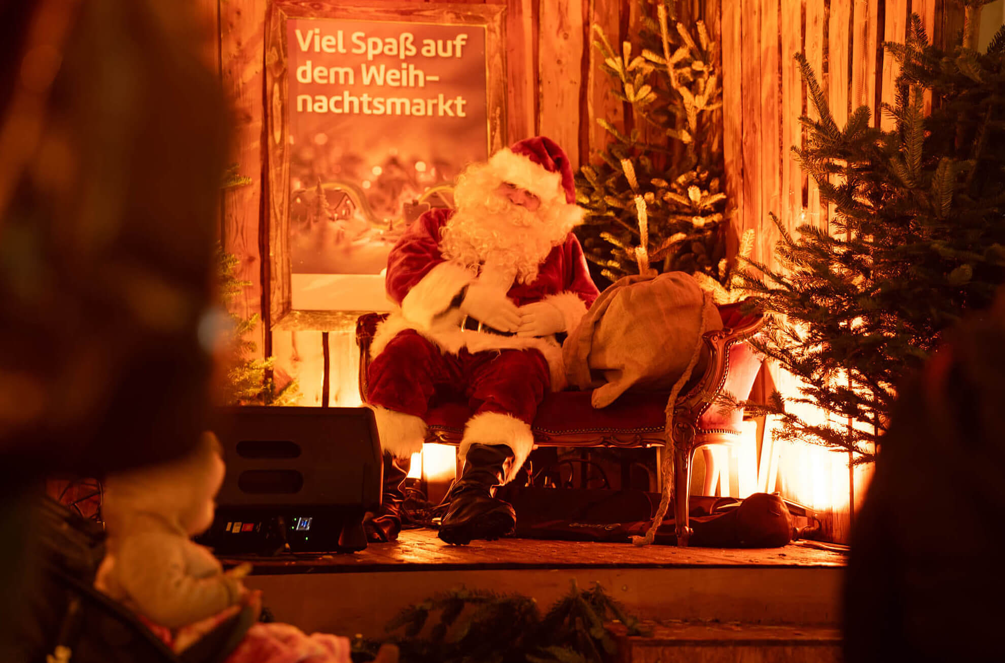 Ein Weihnachtsmann sitzt auf einer Bühne, umgeben von Tannenbäumen.