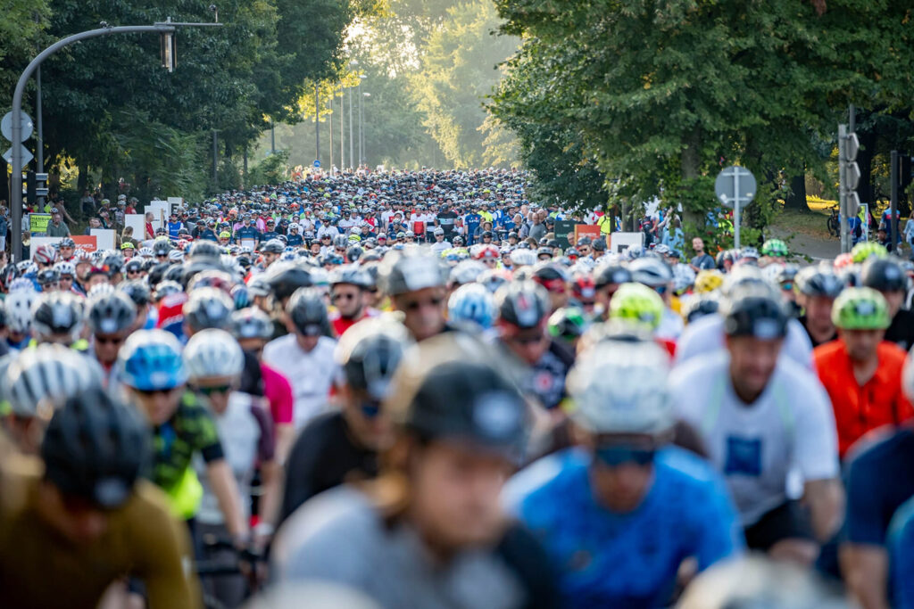 Eine große Anzahl von Radfahrern trägt Helme und fährt auf einer Straße, umgeben von Bäumen.