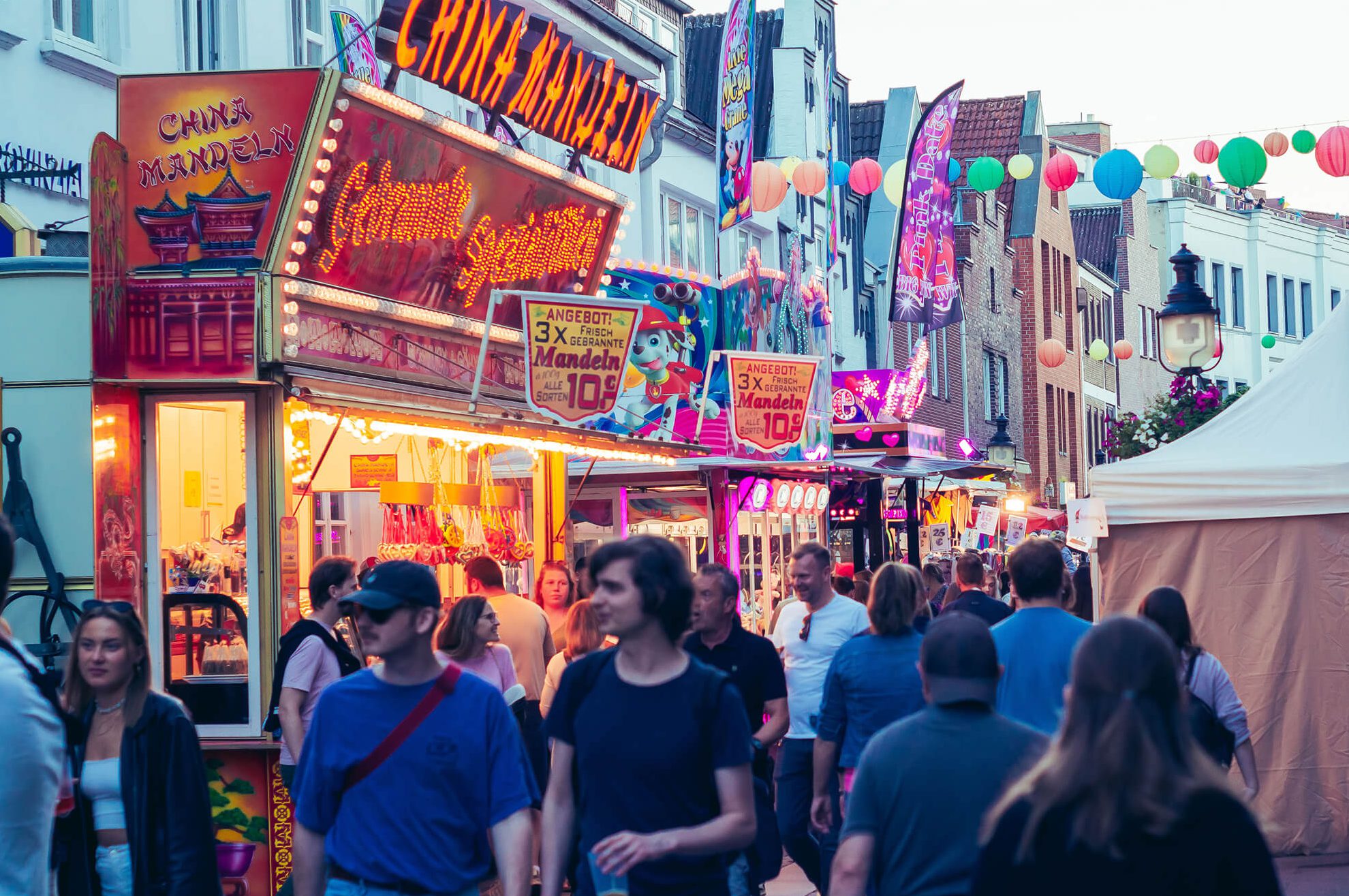 Menschen schlendern entlang einer hell erleuchteten Kirmesstraße mit Verkaufsständen.